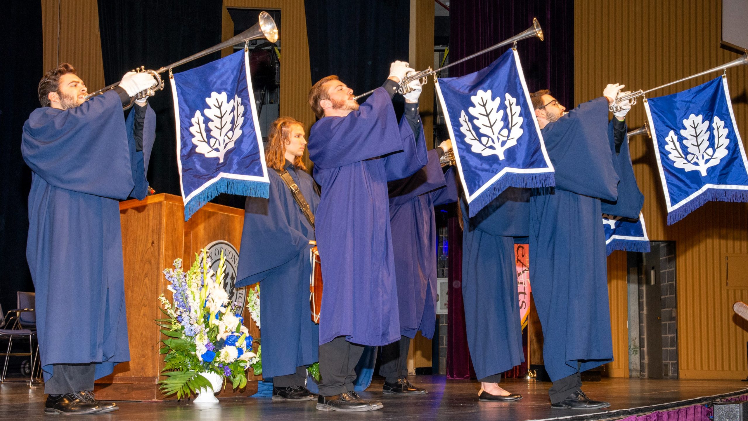 Group of people in academic robes playing herald trumpets with UConn insignia flags on a stage