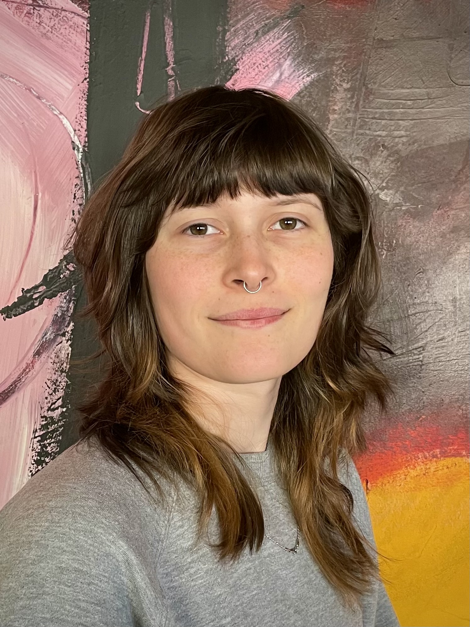 A smiling young woman with shoulder length brown hair and bangs, pierced septum ring, grey shirt, in front of an abstract painting in pink, brown, and yellow tones